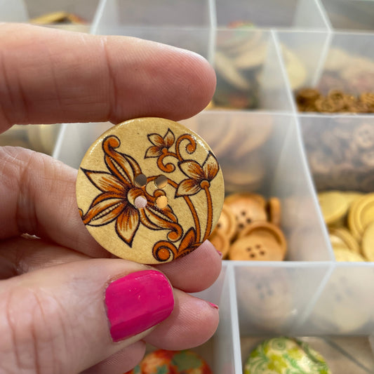 Gold button held by a hand with pink nail polish, against a background of various buttons.