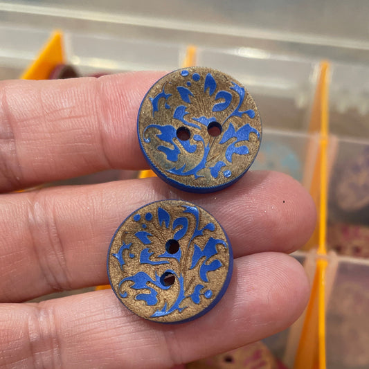 Two decorative buttons held between fingers with a container of buttons in the background.