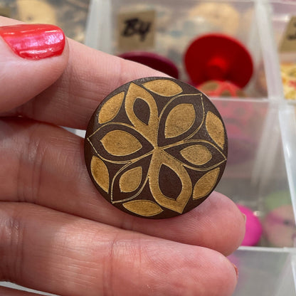 Hand holding a decorative wooden button with a detailed pattern, against a background of various buttons in a container.