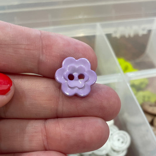 Hand holding a purple flower-shaped button with a background of assorted buttons in a container.