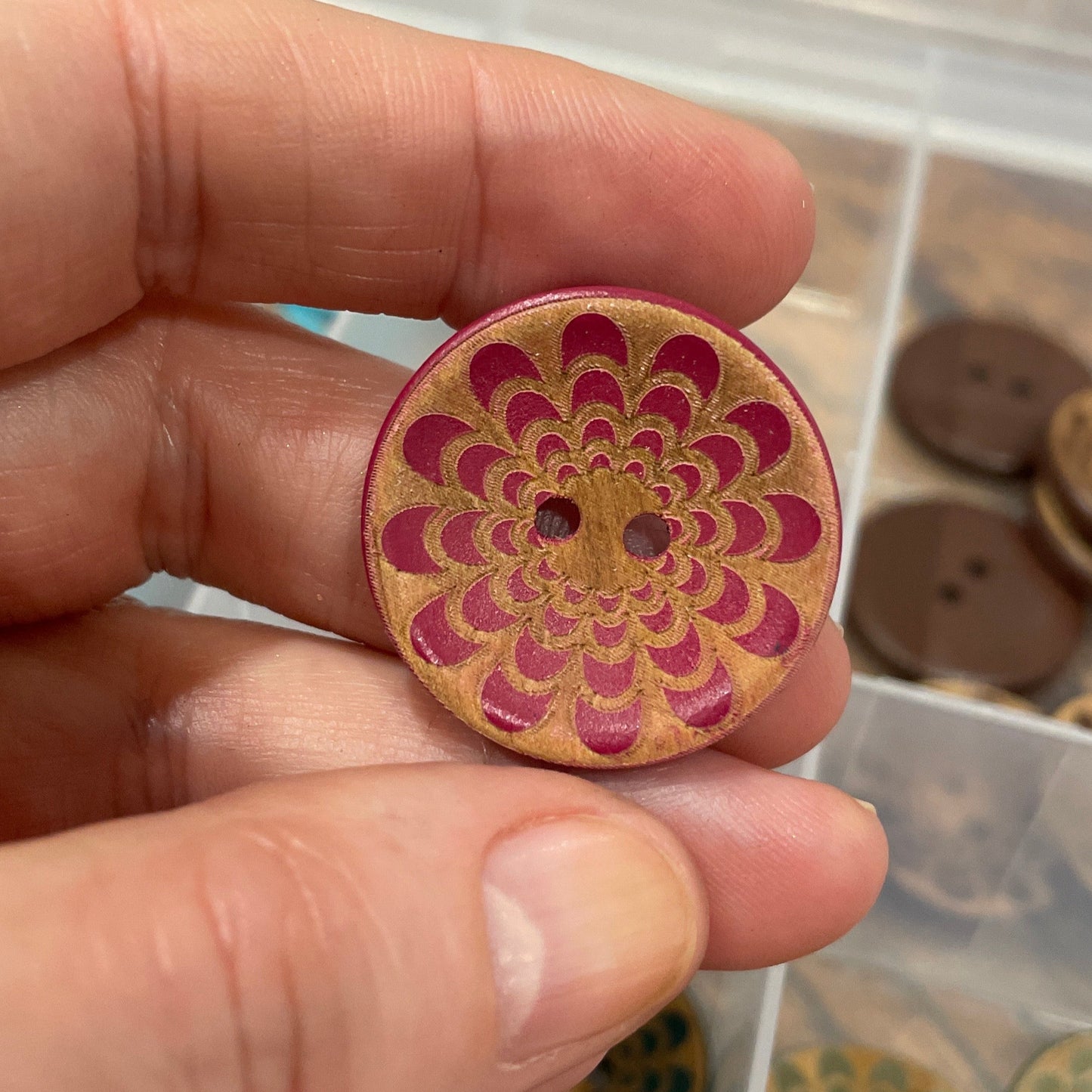 Hand holding a wooden button with a floral pattern, with a container of buttons in the background.