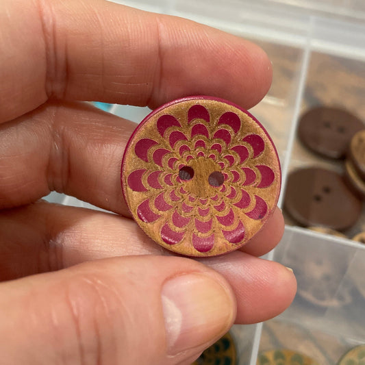 Hand holding a wooden button with a floral pattern, with a container of buttons in the background.
