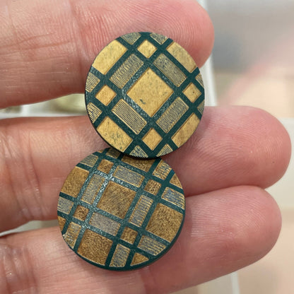 Two round, gold and green patterned buttons held between fingers with a container of buttons in the background.