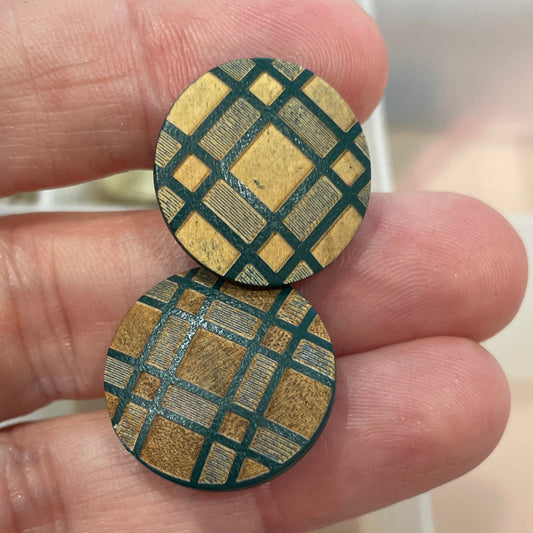 Two round, gold and green patterned buttons held between fingers with a container of buttons in the background.