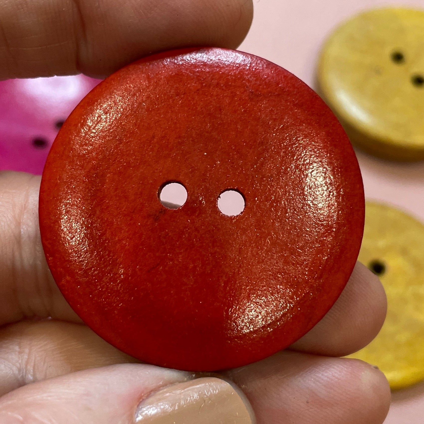 Hand holding a red button with two holes, surrounded by various colored buttons on a pink surface.