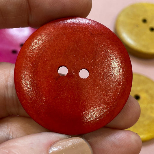 Hand holding a red button with two holes, surrounded by various colored buttons on a pink surface.
