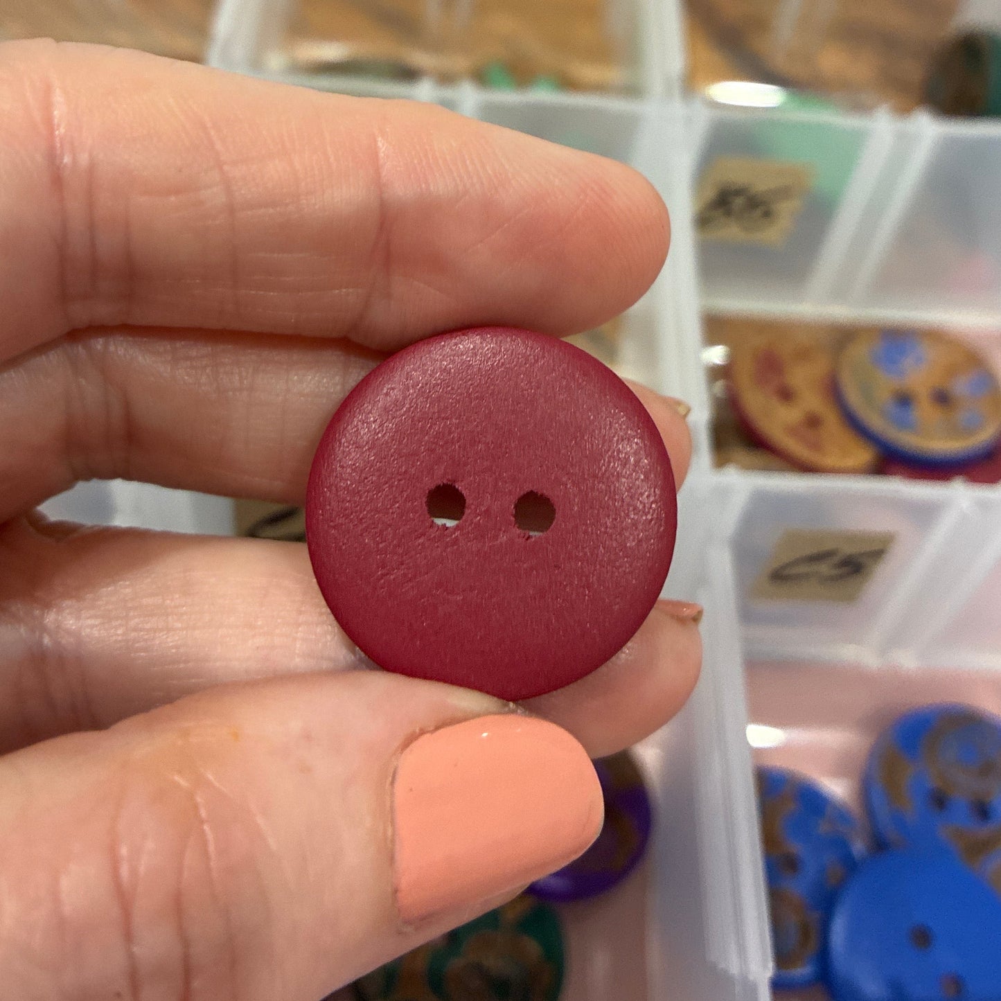 Hand holding a red button with a blurred background of a storage box with buttons.