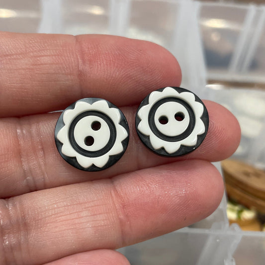 Black and white button-shaped earrings held between fingers with a container of buttons in the background.