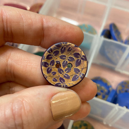 Hand holding a decorative button with intricate design, against a background of a clear container with more buttons.