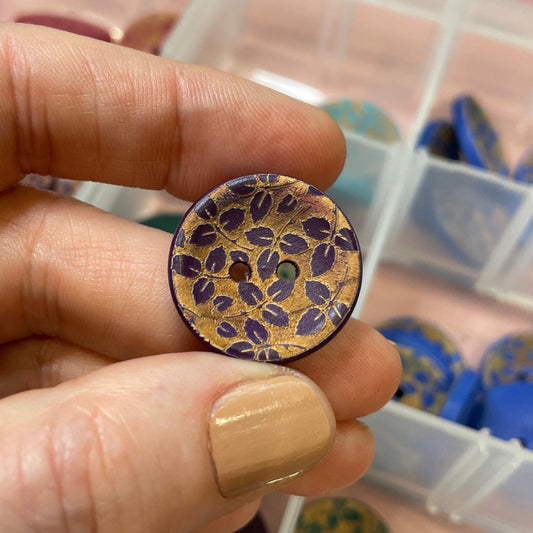 Hand holding a decorative button with intricate design, against a background of a clear container with more buttons.