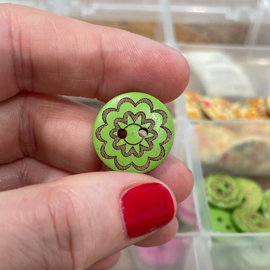 Hand holding a green button with floral design in front of a container of various buttons.
