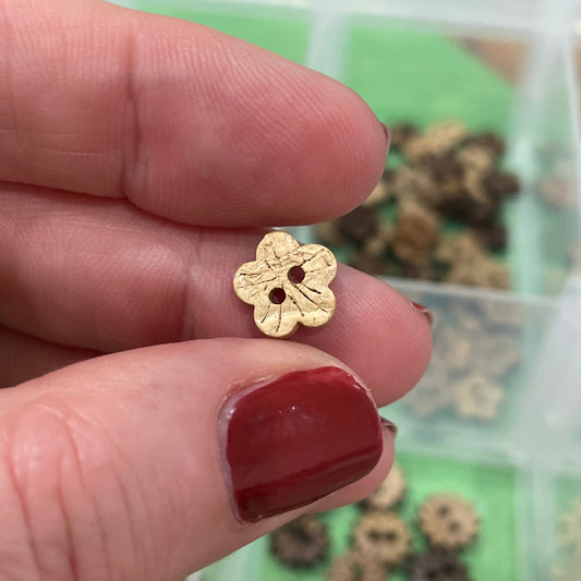 Gold flower-shaped button  held between fingers with a container of decorative items in the background.