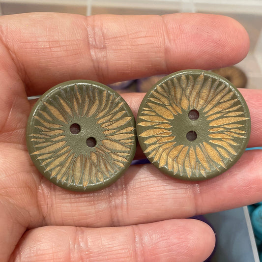 Two bronze buttons held in a hand with a container of assorted buttons in the background.