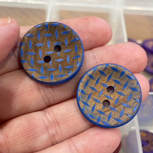 Hand holding two blue patterned buttons with a container of buttons in the background.