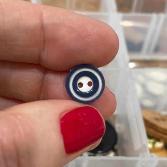 Hand holding a small blue and white button with a blurred background of a button collection.