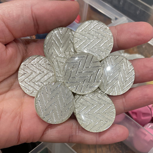 Hand holding silver hexagonal buttons with a container of various colored buttons in the background.