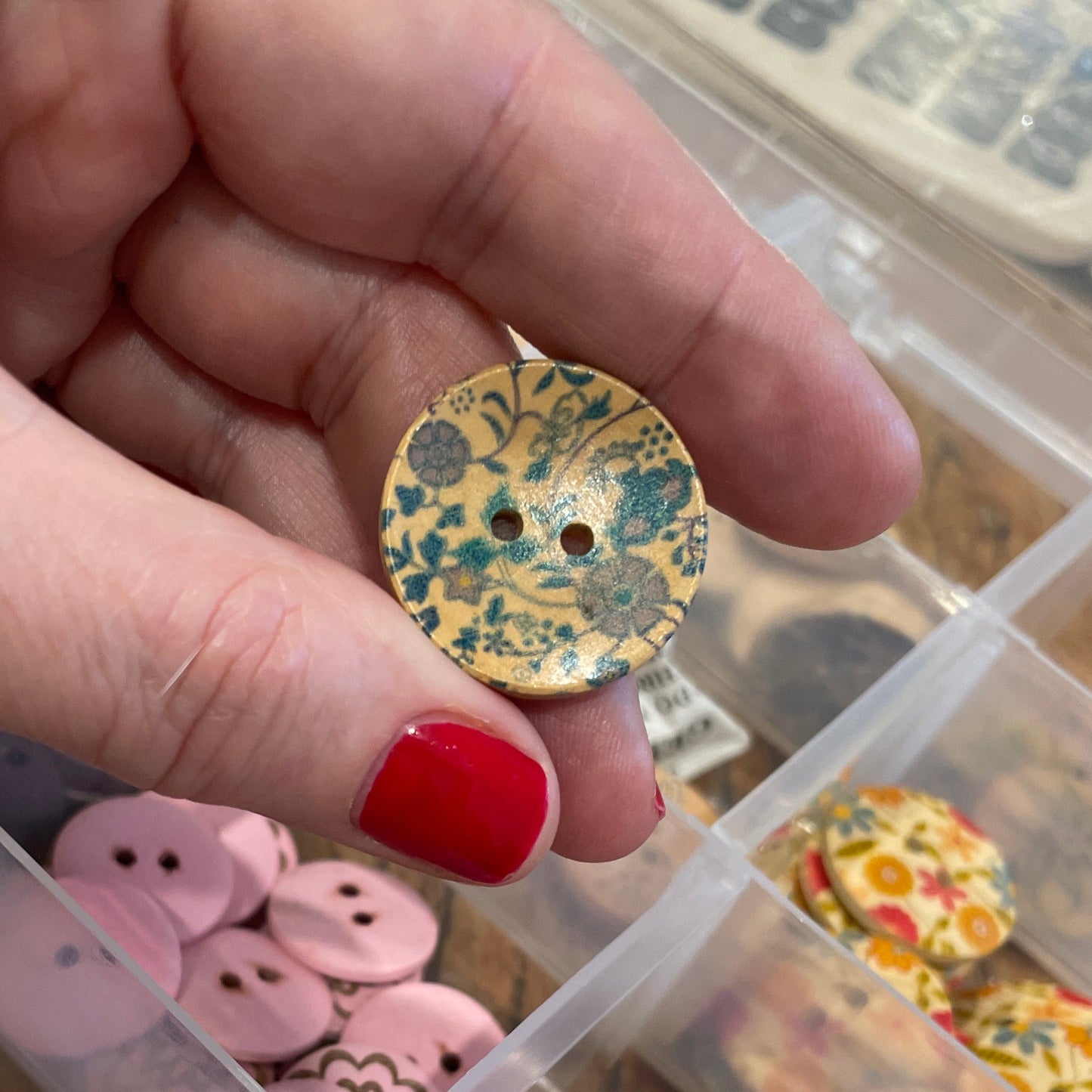 Hand holding a decorative button next to a container of assorted buttons.