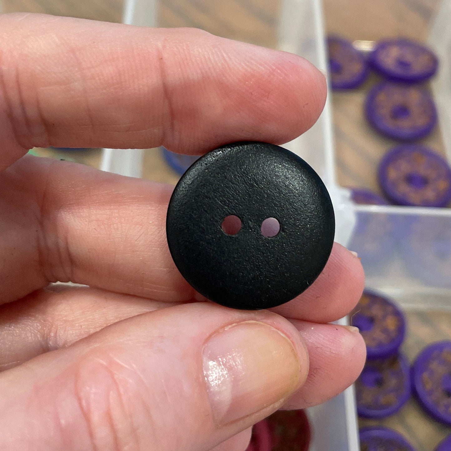Black button held between fingers with a container of buttons in the background