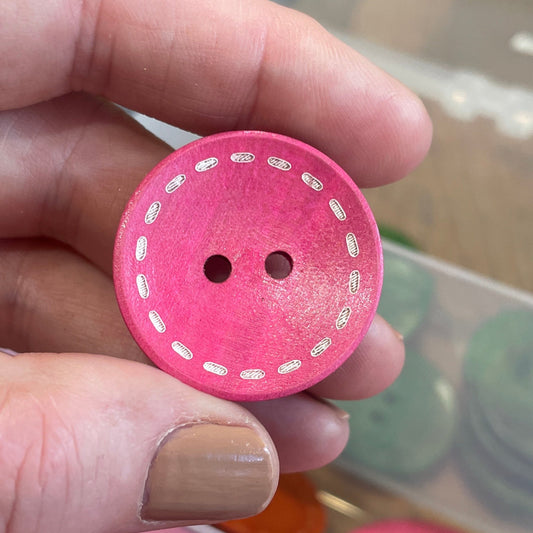 Hand holding a pink button with a patterned edge, with a container of buttons in the background.