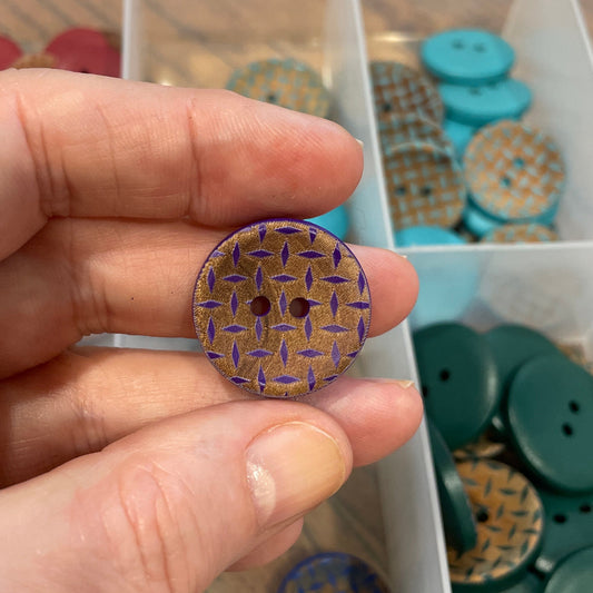 Hand holding a decorative button with a patterned design, against a background of a clear container with various buttons.