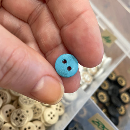 Hand holding a small blue button with a container of various buttons in the background
