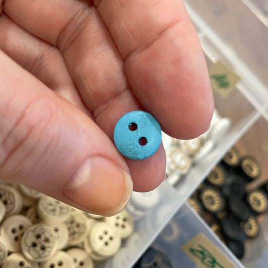Hand holding a small blue button with a container of various buttons in the background