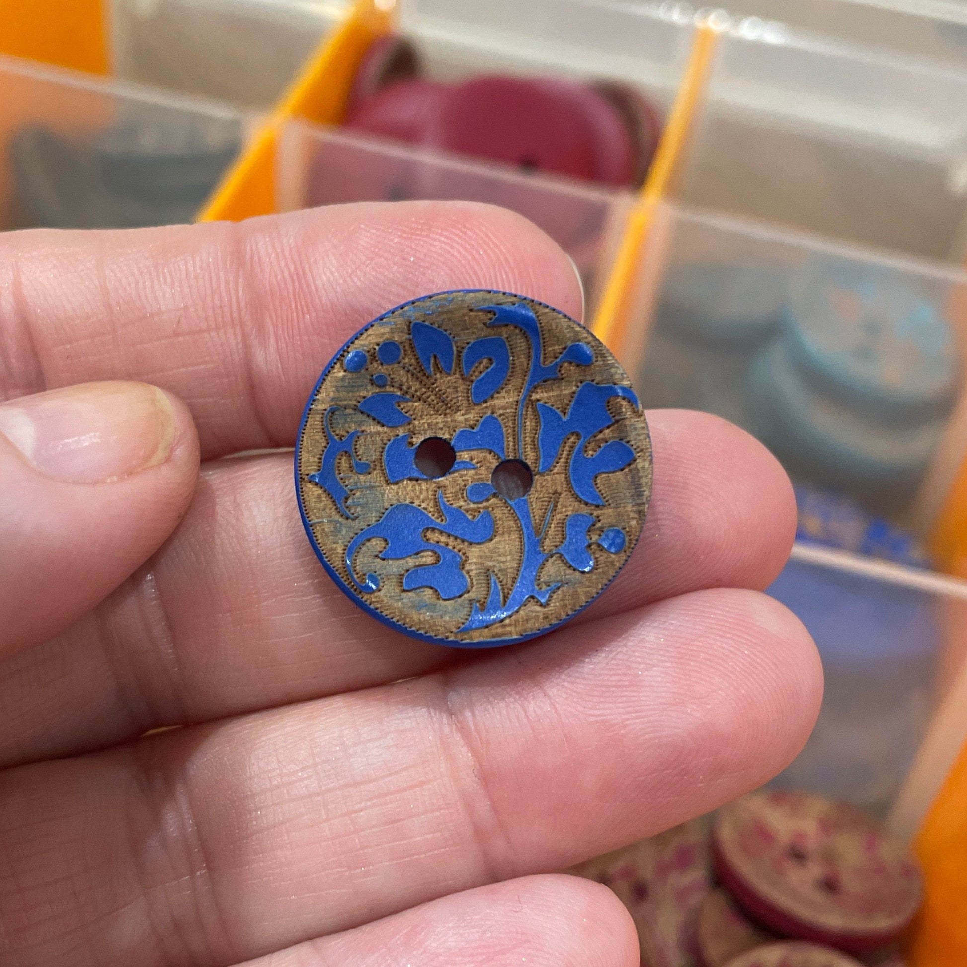 Hand holding a decorative button with blue and gold design in front of a container of buttons.