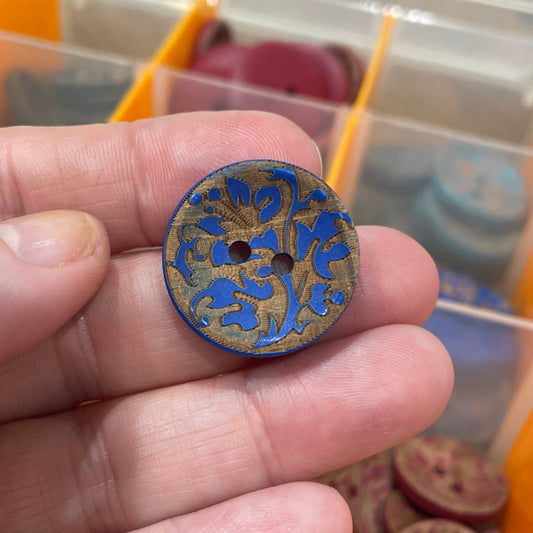 Hand holding a decorative button with blue and gold design in front of a container of buttons.