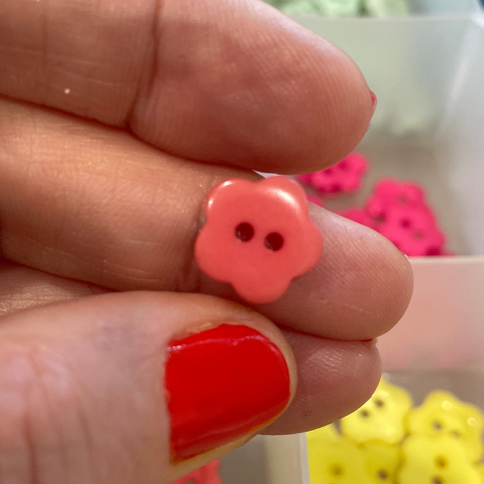 Hand holding a pink flower-shaped button with a container of colorful buttons in the background.