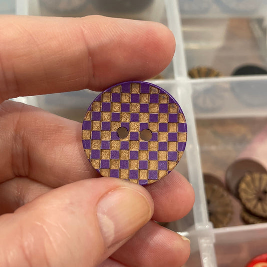 Hand holding a purple and gold checkered button with a container of buttons in the background