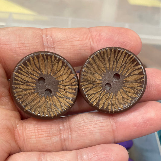 Hand holding two brown decorative buttons with a container of colorful buttons in the background.