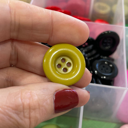 Hand holding a yellow button with a clear container of assorted buttons in the background