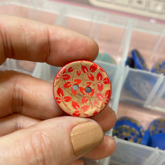 Hand holding a red patterned button with a container of buttons in the background