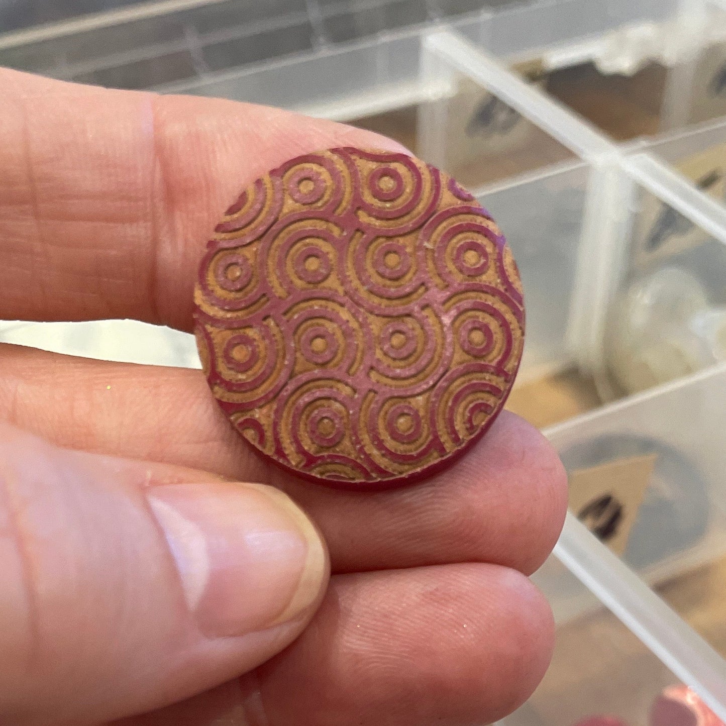 Hand holding a button with intricate patterns in front of an open container of buttons.