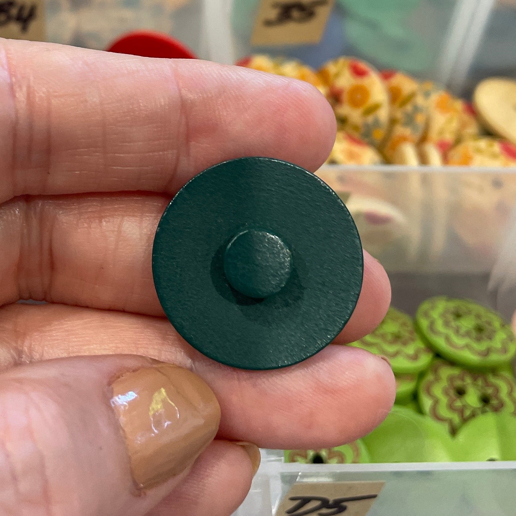 Hand holding a green button with a blurred background of a button collection in a box.