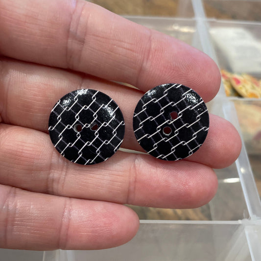 Two black buttons with a textured surface held between fingers, with a clear plastic container in the background.