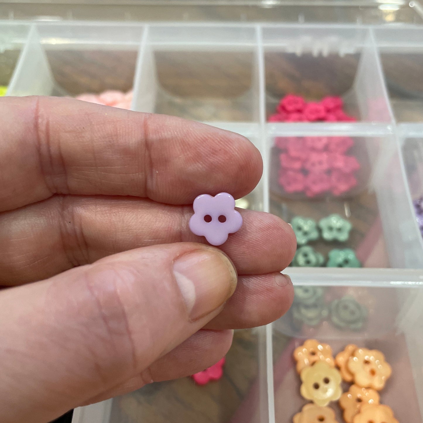 Hand holding a small pink flower-shaped object with a clear container of assorted small objects in the background.