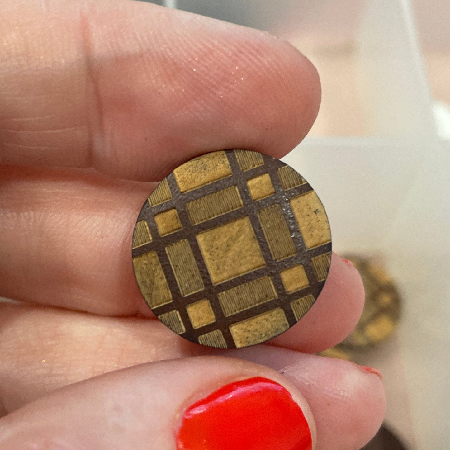 Hand holding a small gold-colored square object with a grid pattern, against a blurred background of buttons.