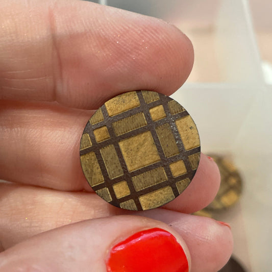 Hand holding a small gold-colored square object with a grid pattern, against a blurred background of buttons.