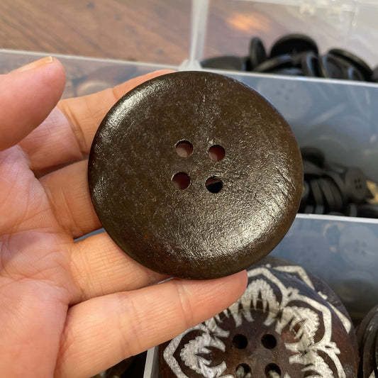Hand holding a dark brown button with three holes, with a display of similar buttons in the background.