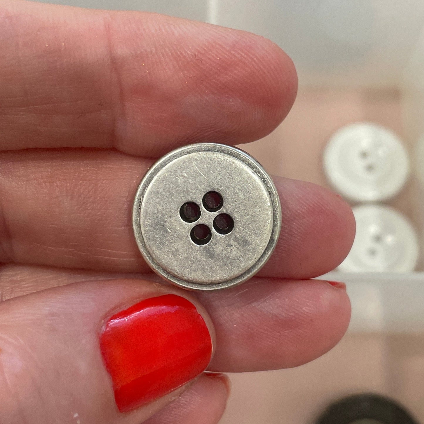 Hand holding a silver button with a clear container of buttons in the background