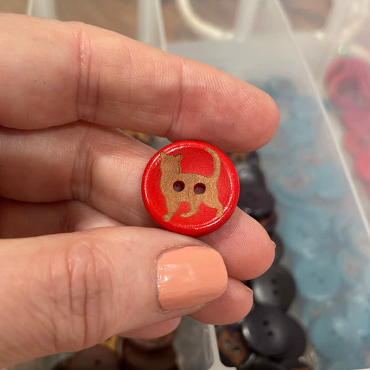 Hand holding a red button with a gold design, against a background of a clear container with various buttons.