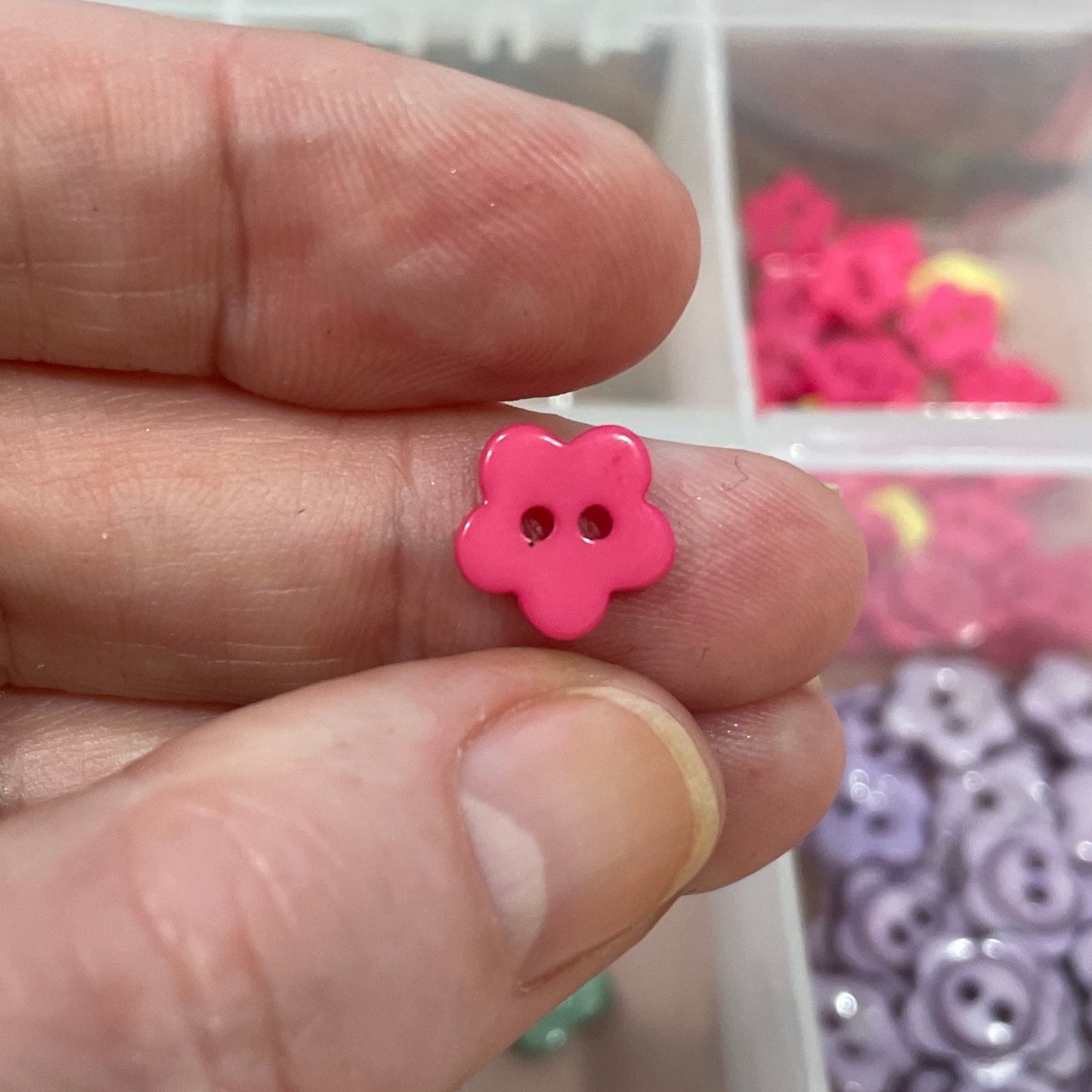 Hand holding a pink flower-shaped button with a container of assorted buttons in the background.