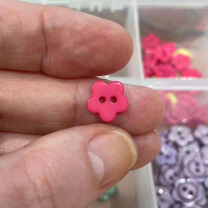 Hand holding a pink flower-shaped button with a container of assorted buttons in the background.