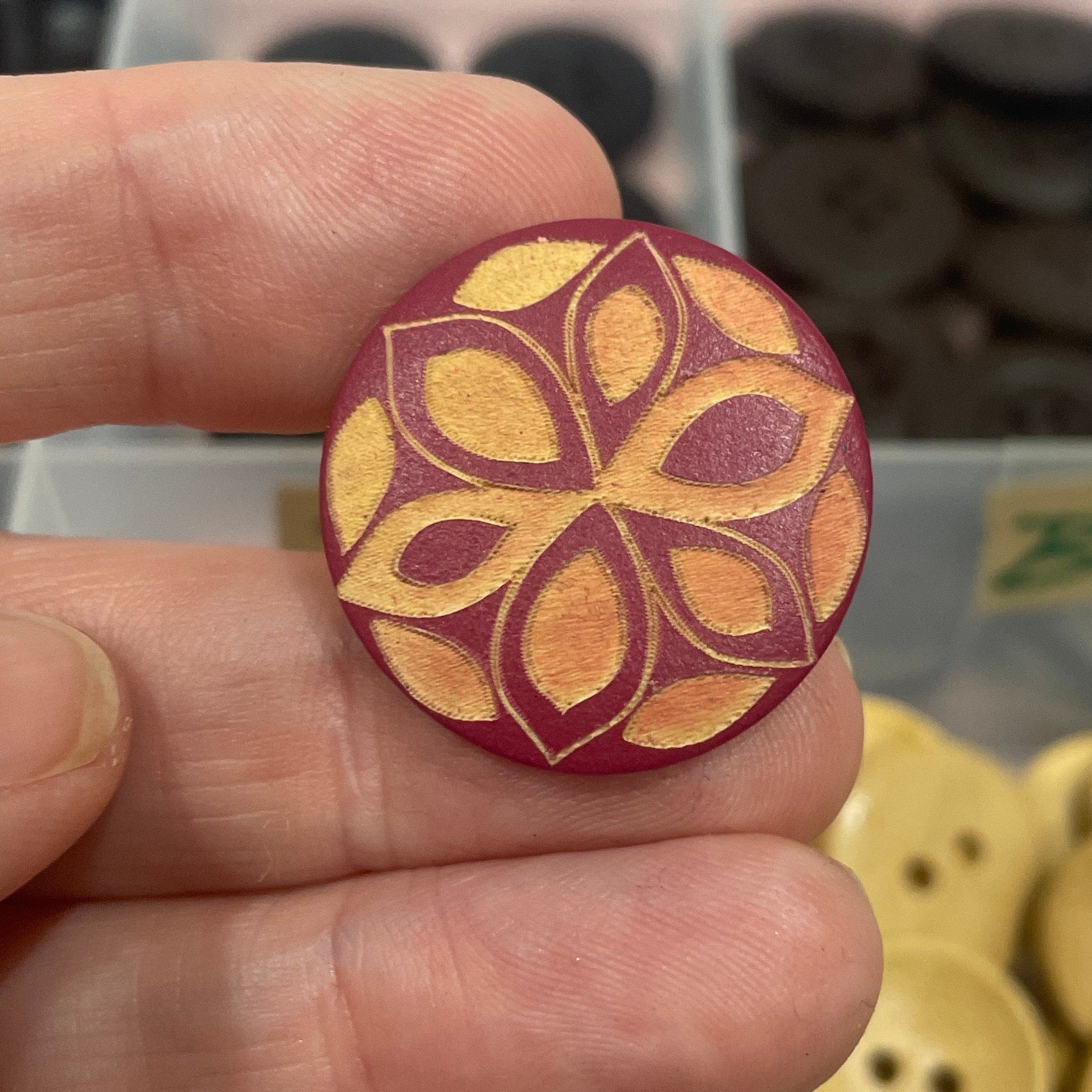 Hand holding a decorative button with a floral pattern, against a background of various buttons.