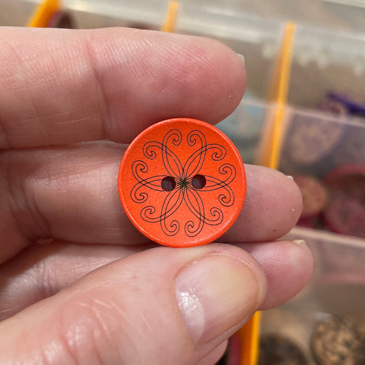 Hand holding an orange button with a decorative pattern against a background of other buttons in a container.