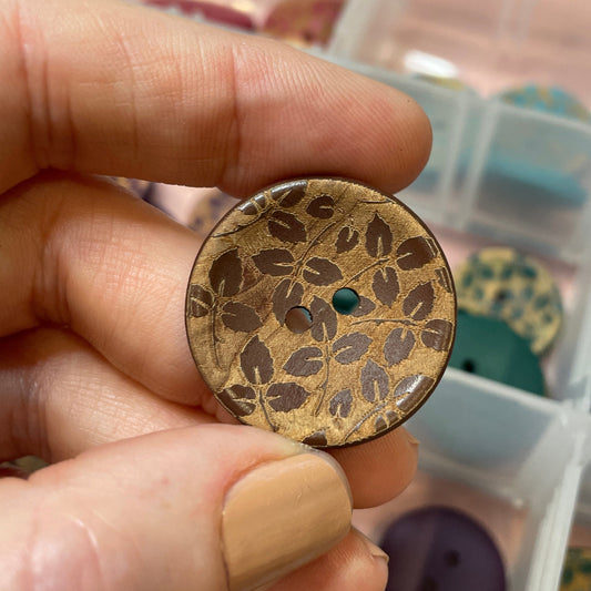 Hand holding a decorative button with a pattern, with a container of buttons in the background.