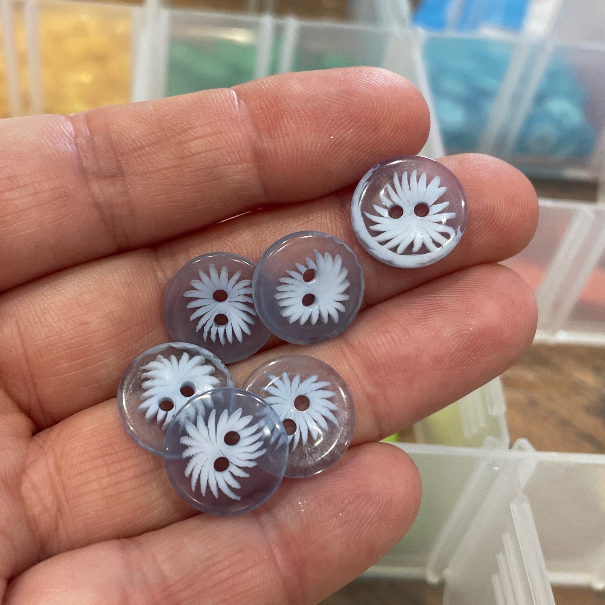Hand holding five clear buttons with white floral designs against a blurred background of button containers.