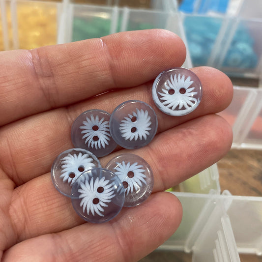 Hand holding five clear buttons with white floral designs against a blurred background of button containers.
