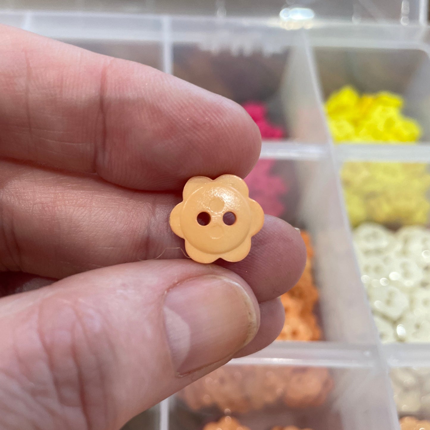 Hand holding a small yellow flower-shaped object with buttons, against a background of similar objects in a container.